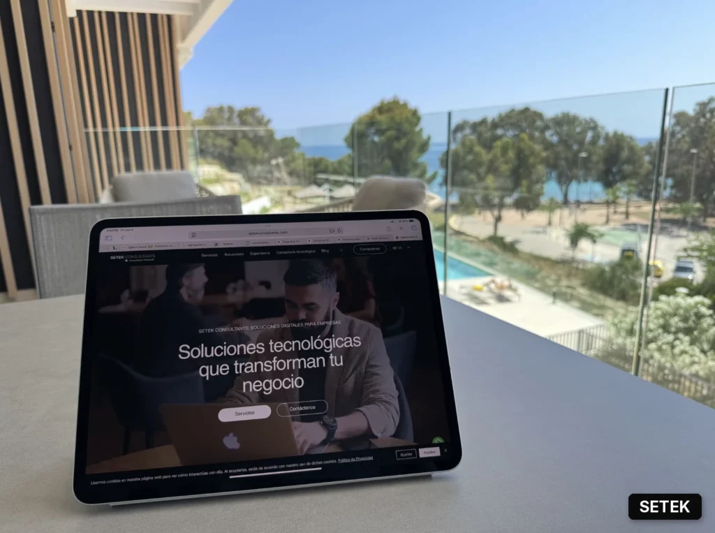 Person working remotely on a MacBook and tablet, showing Setek Consultants' mobile device management dashboard, overlooking coastline